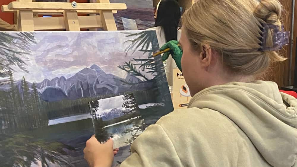 Student painting a serene mountain landscape in an art class, with a focus on learning acrylic techniques