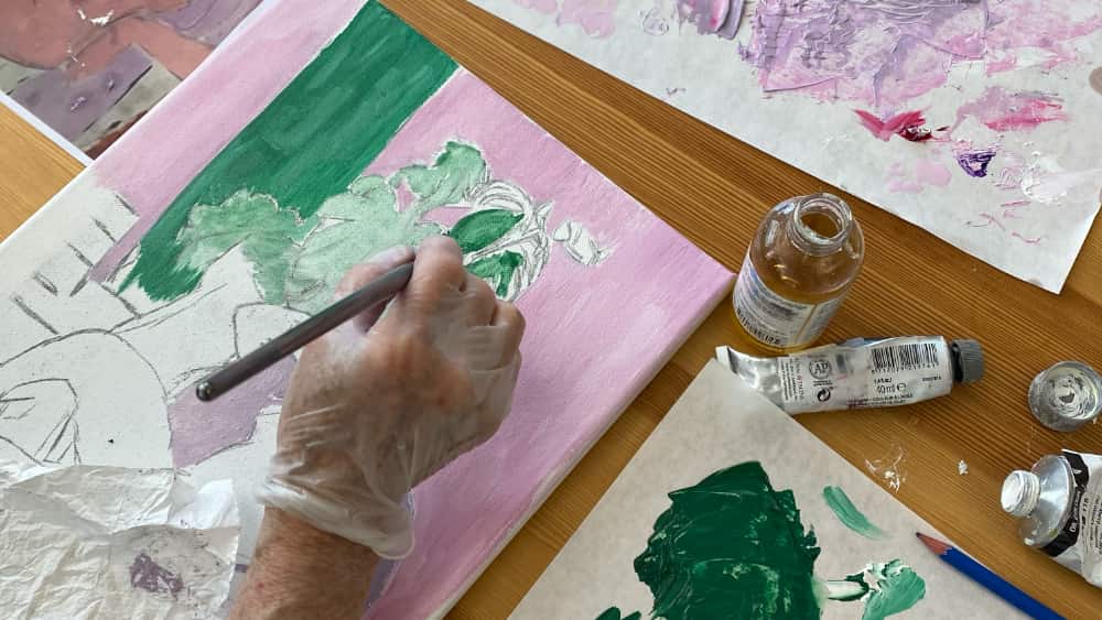 A close-up of a student's hand painting a green floral pattern on a pink canvas, with acrylic paint tubes and a mixing palette in view