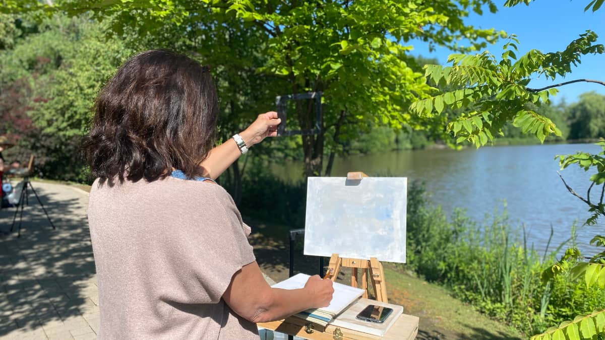 Artist practicing plein air painting by a lake, capturing the scene on a clear canvas, with lush greenery and bright daylight enhancing the creative ambiance