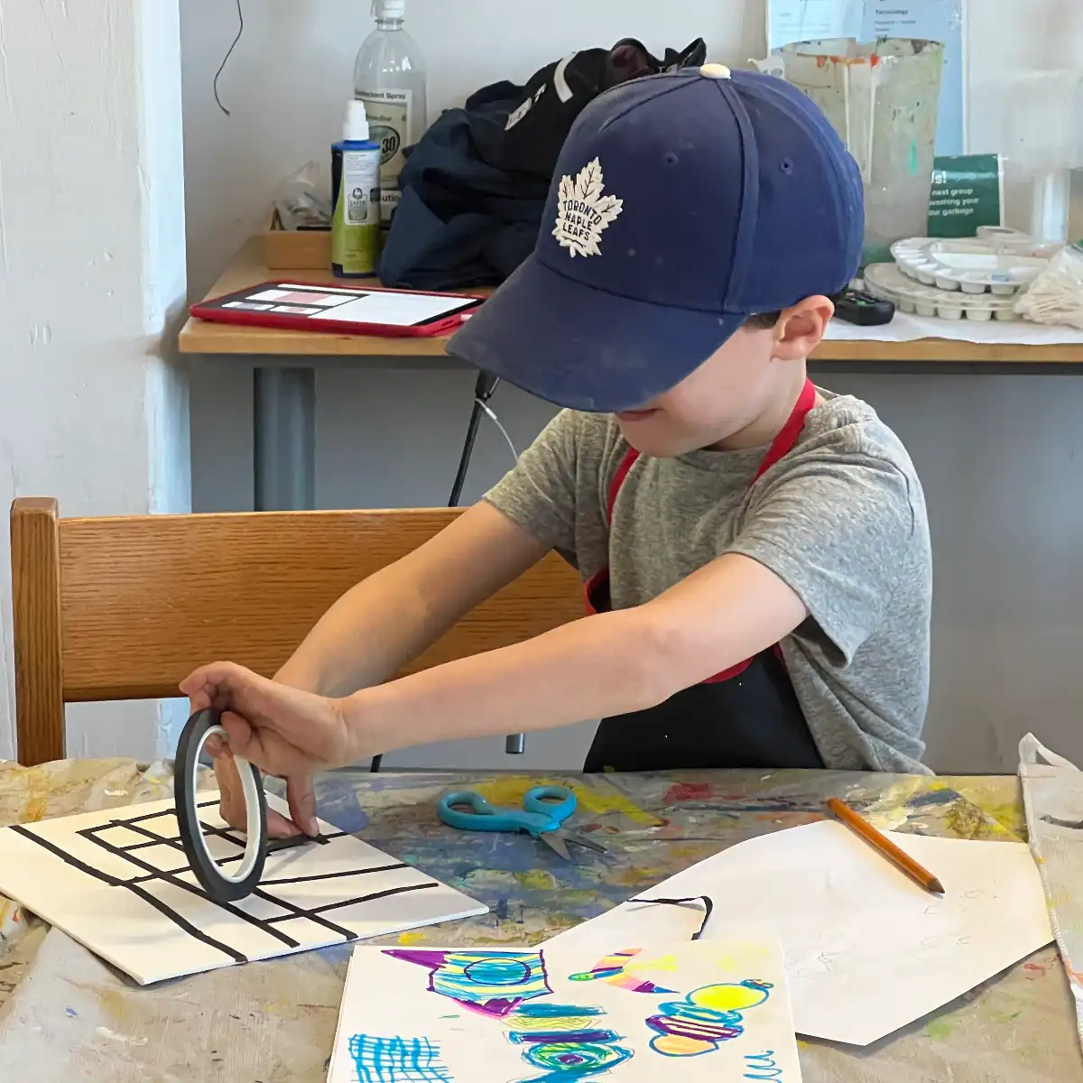 A child wearing a blue baseball cap applies black tape to a white board to create a grid pattern next to colorful marker illustrations Drawing, Painting and Making - Kids at Avenue Road Arts School — Winter 2026