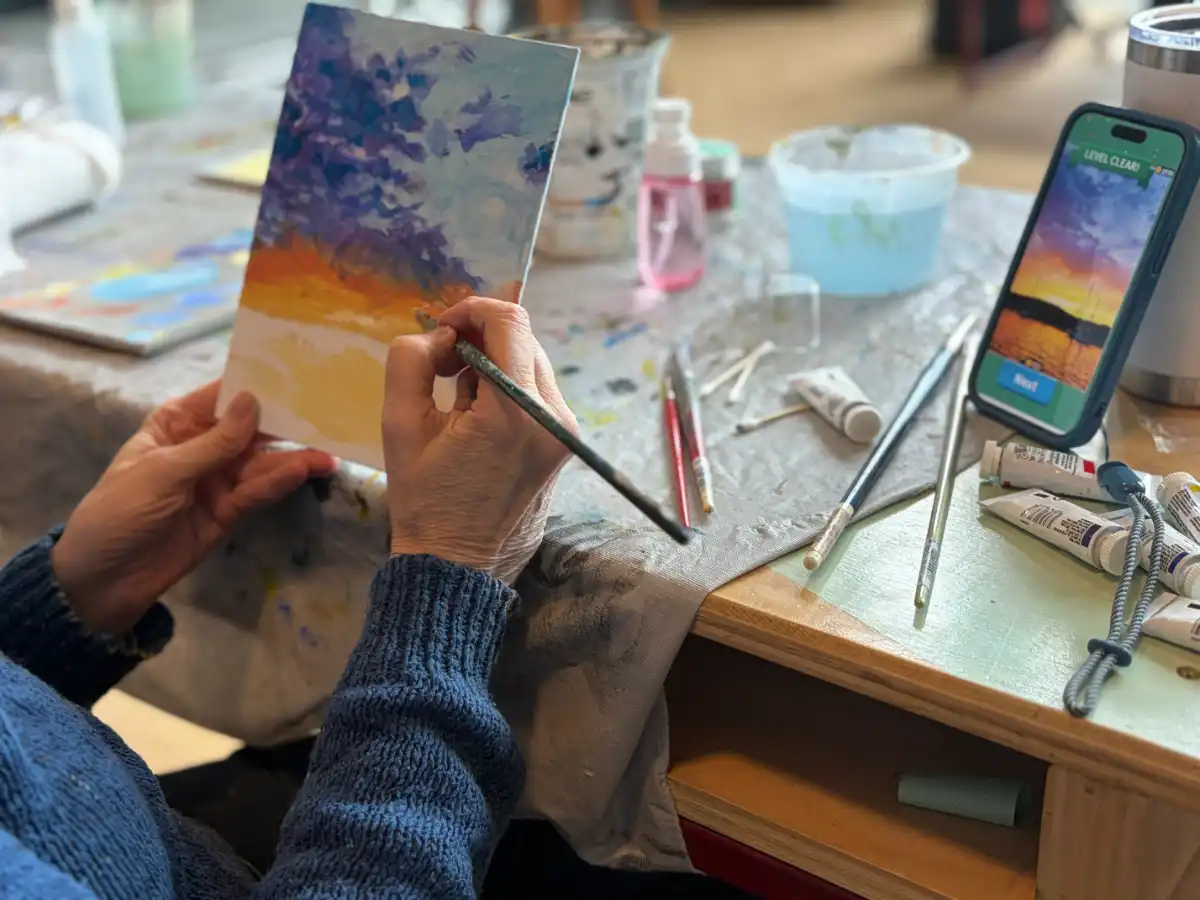 A hand in a blue sweater applies orange and yellow paint to a small landscape board next to gouache tubes and a smartphone displaying a reference photo Gouache Immersion at Avenue Road Arts School — Winter 2026