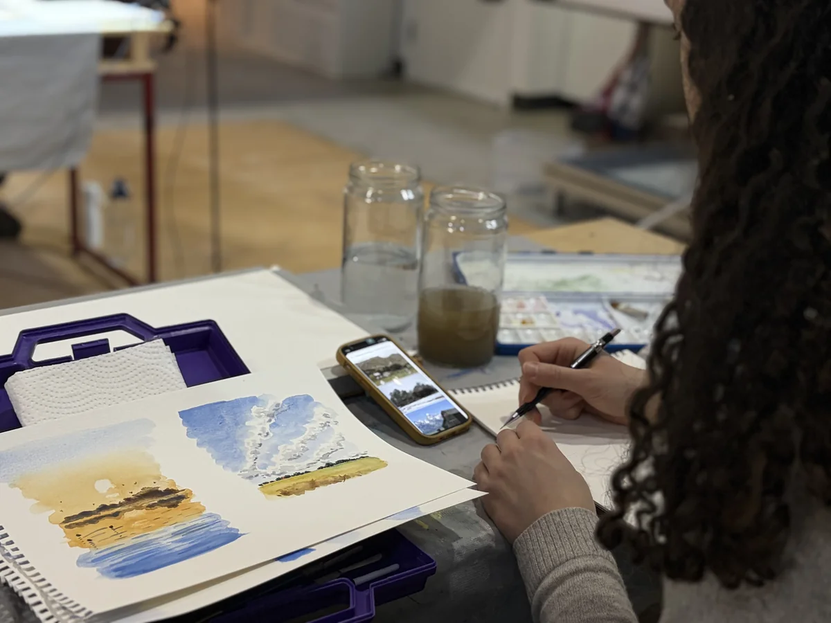 Student painting a golden autumn landscape in watercolour, working from a photo reference in a bright art studio