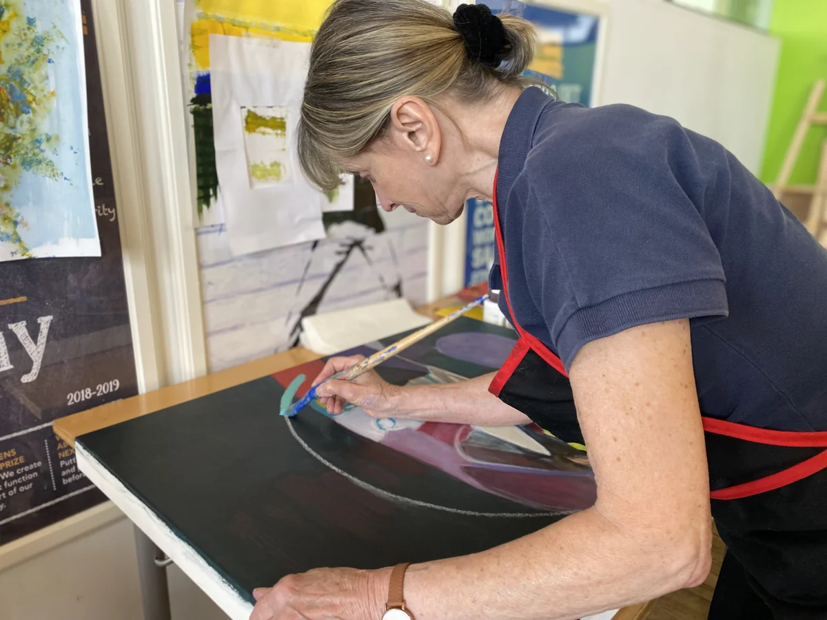 Artist in an apron blending soft pastel on a large canvas at an easel in a colourful studio