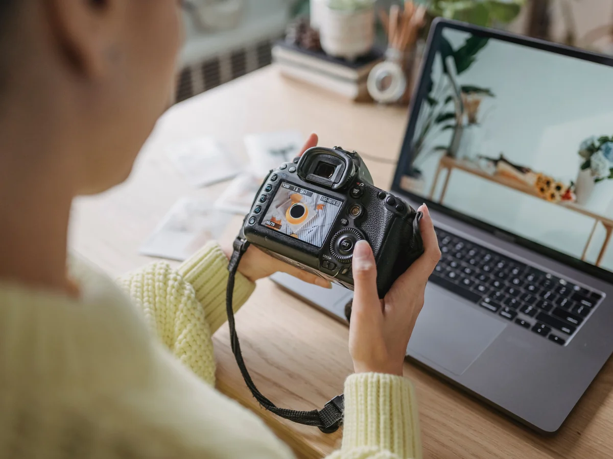 Person reviewing a photograph on the back of a DSLR camera beside a laptop on a wooden desk
