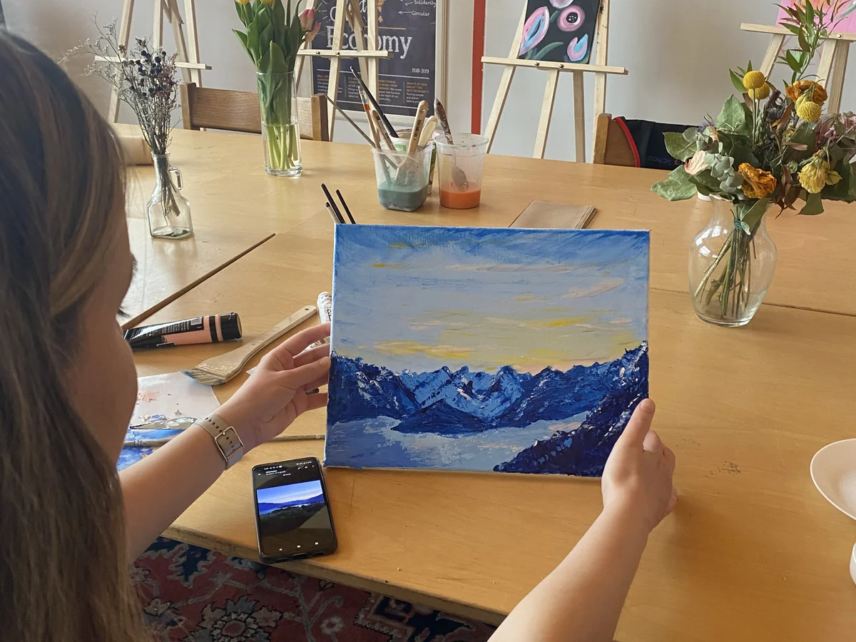 Student holding a finished acrylic painting of snow-capped mountains above a lake in a flower-filled studio