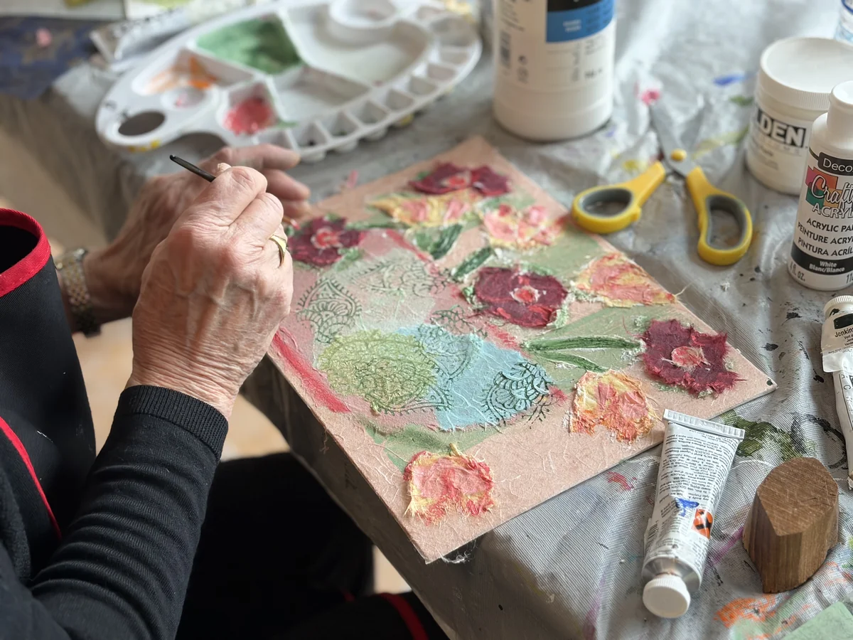 Artist's hands painting a textured floral mixed-media collage with acrylic and patterned paper