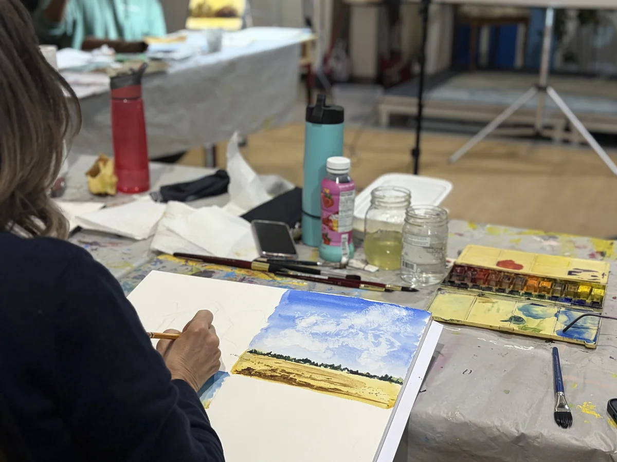 Student painting a watercolour landscape of fields and sky in a busy communal art studio